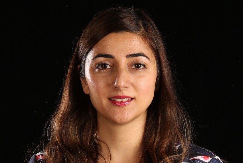 A woman with long hair wearing a blue shirt with splashes of bright pink stands against a black background, exuding a vibrant yet elegant presence.