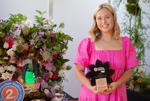 Chantal Williams, Second Prize Winner of Chrysco Intermediate Floristry Competition, in a pink dress holding an award with her floral design on display.