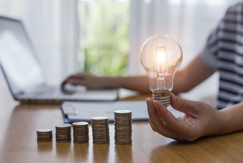 Stacks of coins arranged on a desk and a person holding a lit lightbulb while typing on a laptop