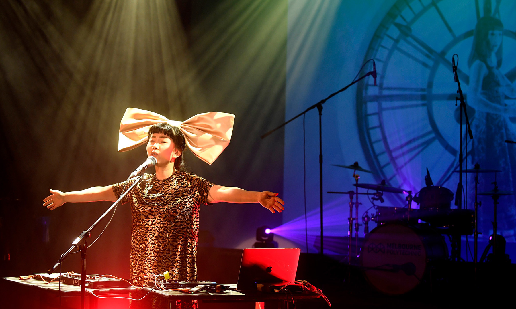 Person performing on stage with a microphone, computer, and MIDI controllers