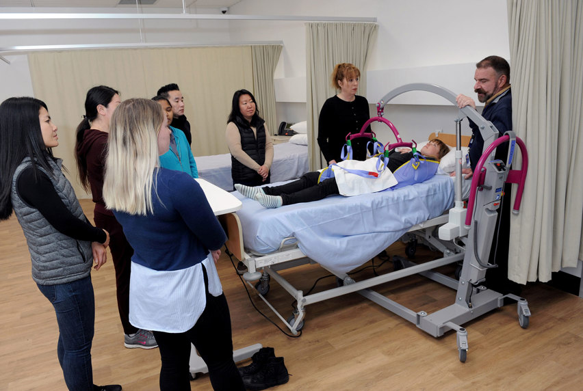 An image of a group of Aged Care students gathered around a patient lying in a hospital bed with a teacher showing how to use a patient hoist.