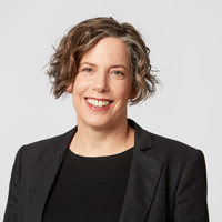 A lady with short brown hair, wearing a black top and blazer smiles at the camera.