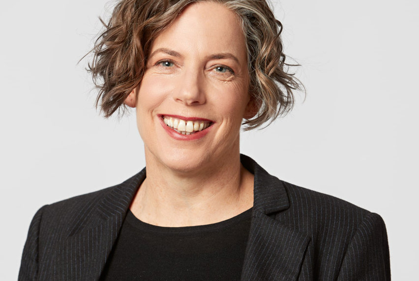 A lady with short brown hair, wearing a black top and blazer smiles at the camera.