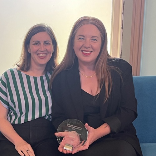 Two women sitting together on a blue couch, smiling at the camera holding a glass award trophy, representing their teamwork and recognition of achievement.