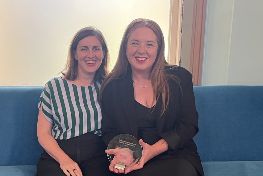 Two women sitting together on a blue couch, smiling at the camera holding a glass award trophy, representing their teamwork and recognition of achievement.