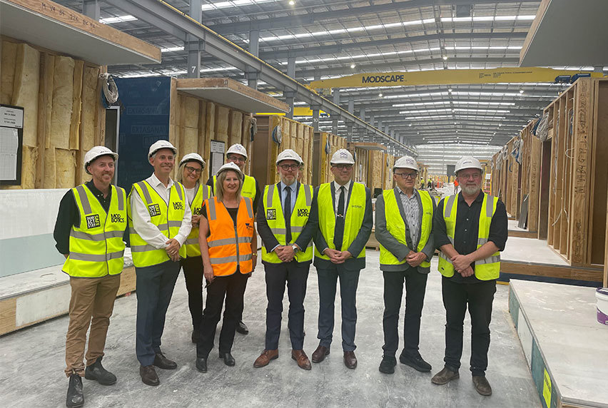 A group of men and a lady wearing hard hats and vests stands together inside a building, engaged in discussion or planning.