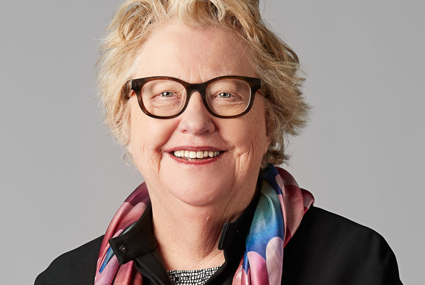 An older lady with short blonde hair smiling at the camera wearing brown glasses and a pink, blue and orange scarf