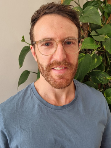 Hayden Park in a light blue t-shirt with glasses, sporting a trimmed beard. A green indoor plant is visible in the background.