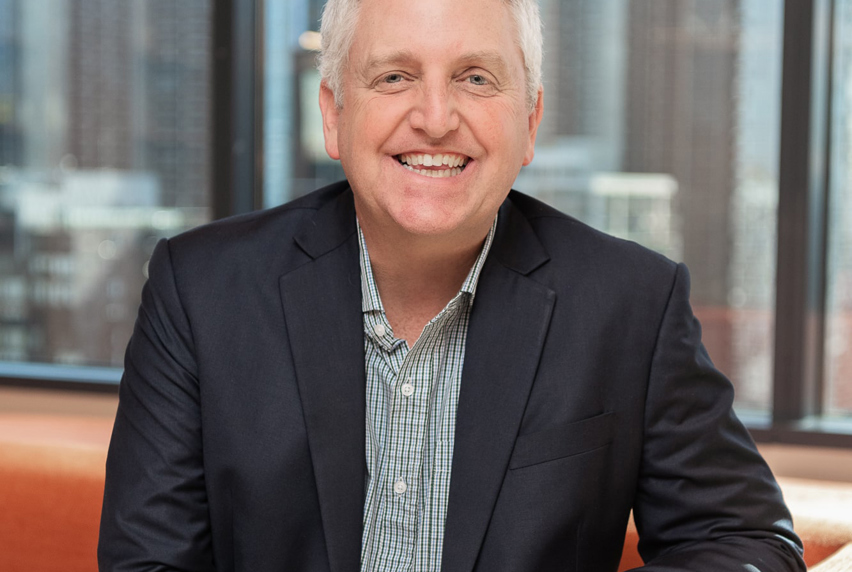 A cheerful businessman wearing a navy suit, smiles at the camera, in front of a city skyline background