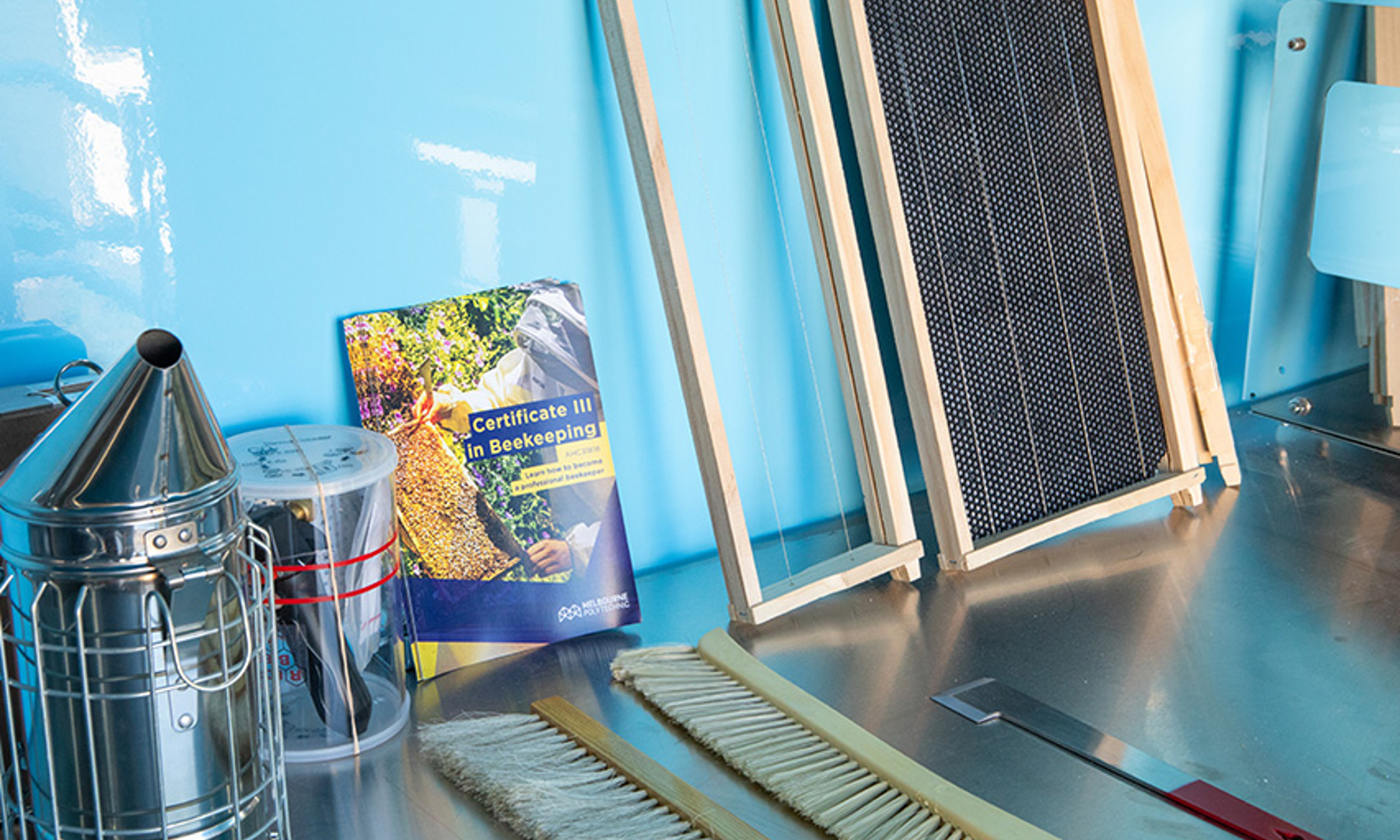 Beekeeping smoker, brush, beekeeping tool, and frame placed alongside a Certificate III in Beekeeping AHC31818 information booklet.