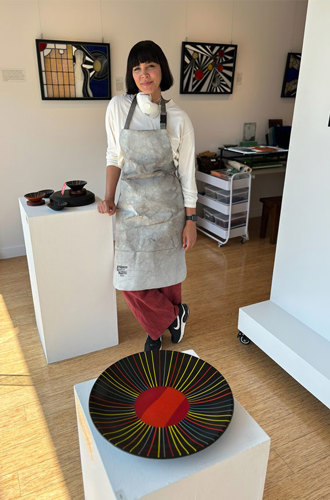 A woman in an apron poses in front of a vibrant glass pieces within a studio.