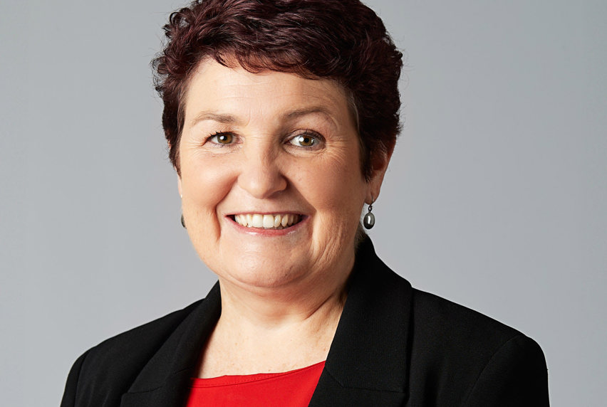 A lady with short dark brown hair, wearing a red top and black blazer smiles at the camera in front of a grey background 