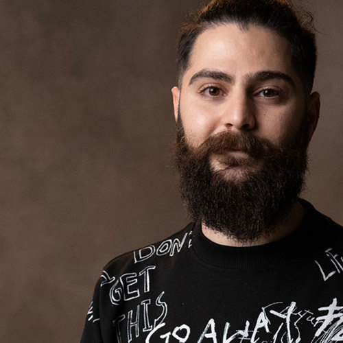 Sam Koraeel smiling and wearing a black t-shirt with expressive text with a neutral background.