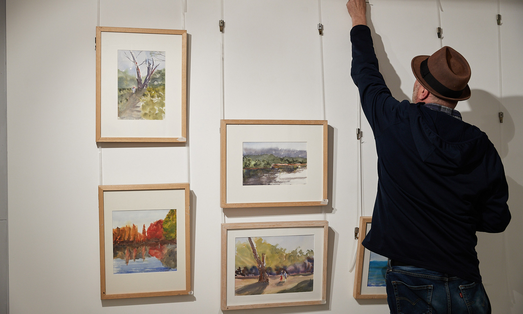 A man wearing a trilby hat adjusting images hung in frames on the wall.