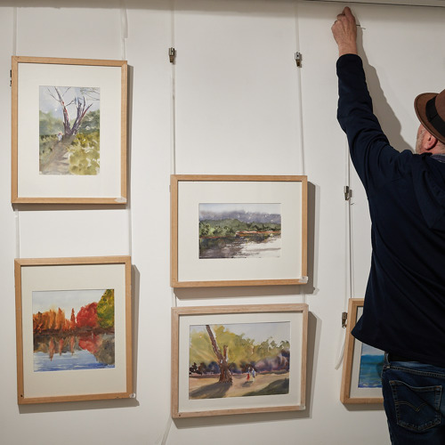 A man wearing a trilby hat adjusting images hung in frames on the wall.