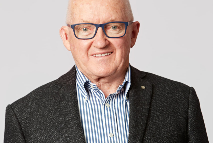 An older man wearing a white and blue stripped shirt, with a charcoal colour blazer and blue glasses smiles at the camera.