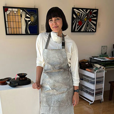 A woman in an apron poses in front of a vibrant glass pieces within a studio.