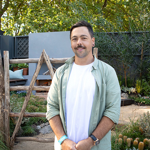 Leigh Hudson stands in a garden that features wooden decor, banksias, and other native plants. He wears a white t-shirt beneath a pale green shirt.