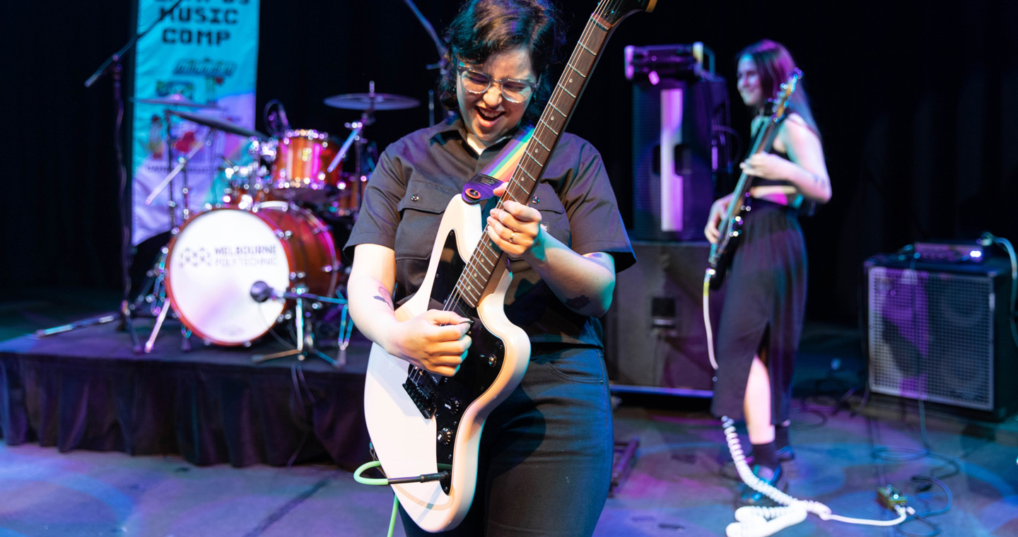 A passionate guitar player on stage at a music competition, joyfully performing with a white electric guitar, with a drummer and another guitarist in the background, under vibrant stage lighting.