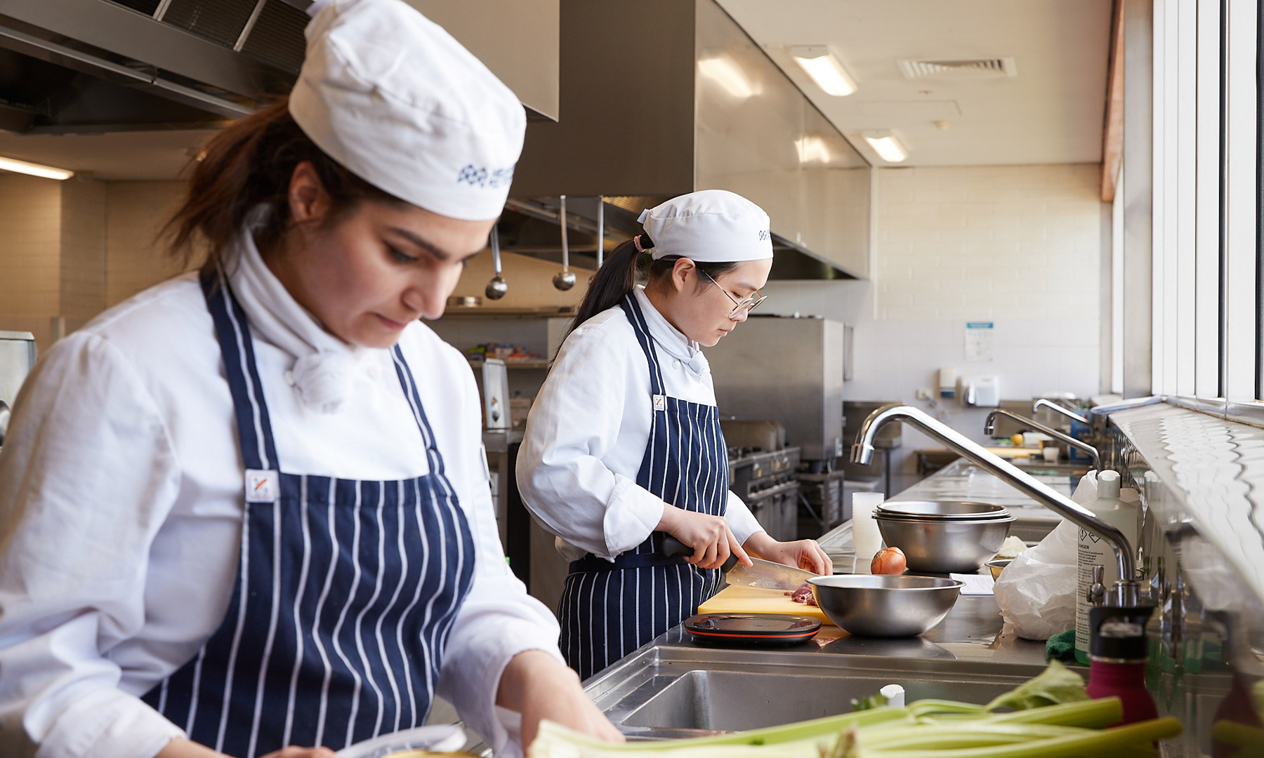 Two hospitality students, in white cook's shirts and navy and white striped aprons, don Melbourne Polytechnic cook's caps while engaged in the process of food preparation.